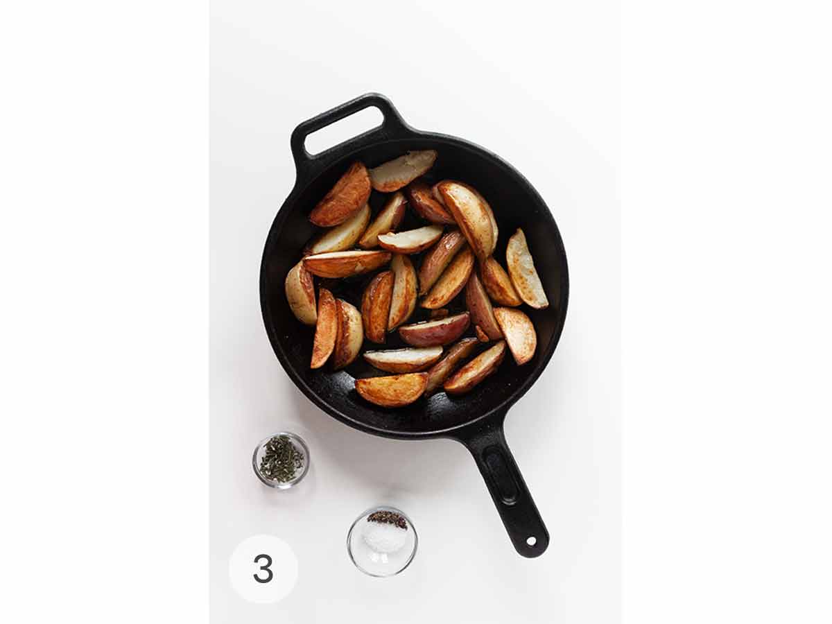 A skillet of roasted grilled potatoes with a dish of chopped rosemary and a dish of salt and pepper beside it.
