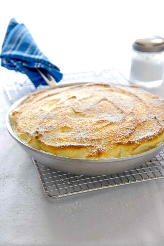 A skillet lemon souffle made entirely in a frying pan, sitting on a wire rack, blue cloth wrapped around the handle.
