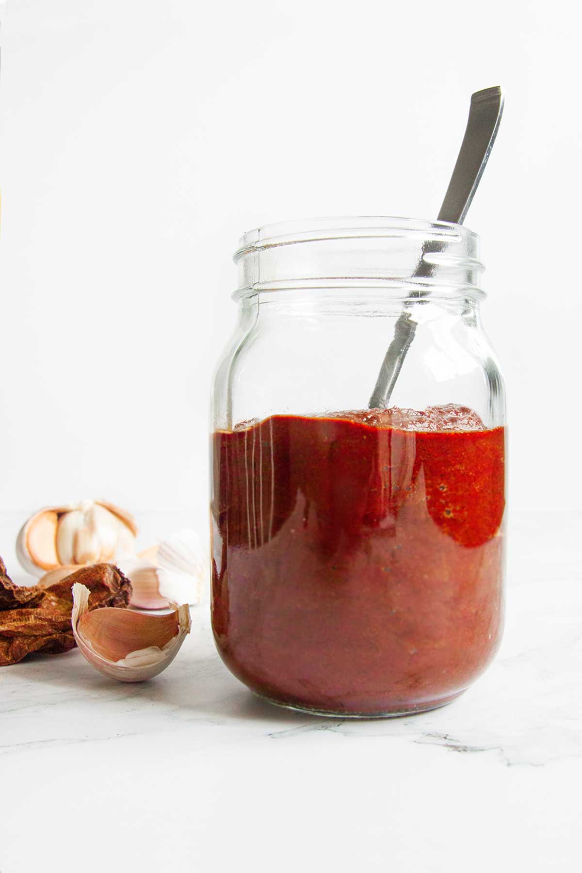 A jar of homemade adobo sauce with a spoon resting inside and garlic cloves and dried chipotles on the side.