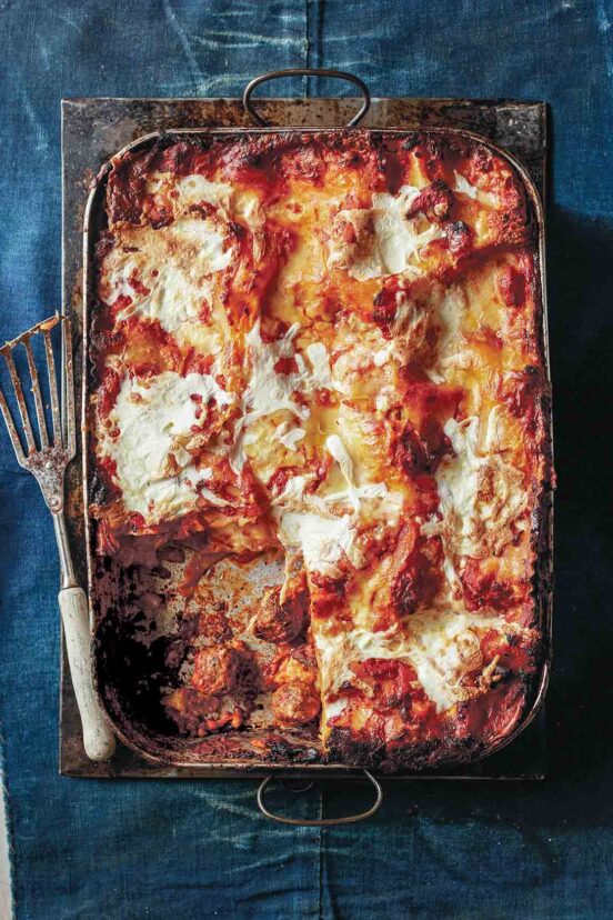 A rectangular baking dish filled with meatball lasagna with one piece missing and a metal spatula on the side.