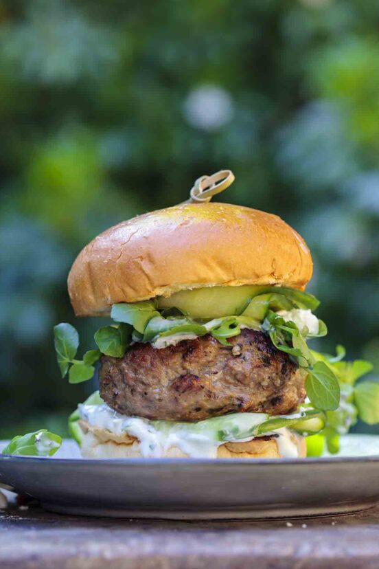 A grilled lamb burger topped with greens and pickles with some tzatziki dripping out.