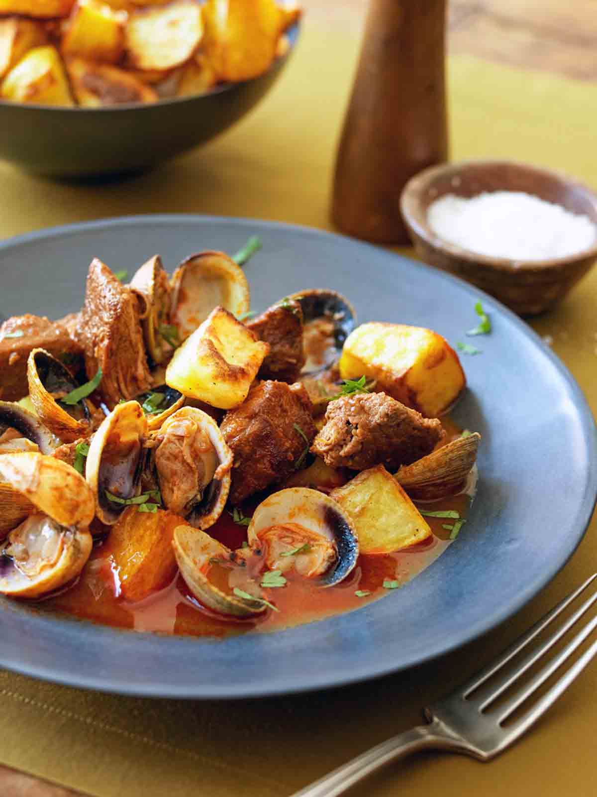 A blue bowl filled with pork and clams with a bowl of potatoes and a dish of salt in the background.