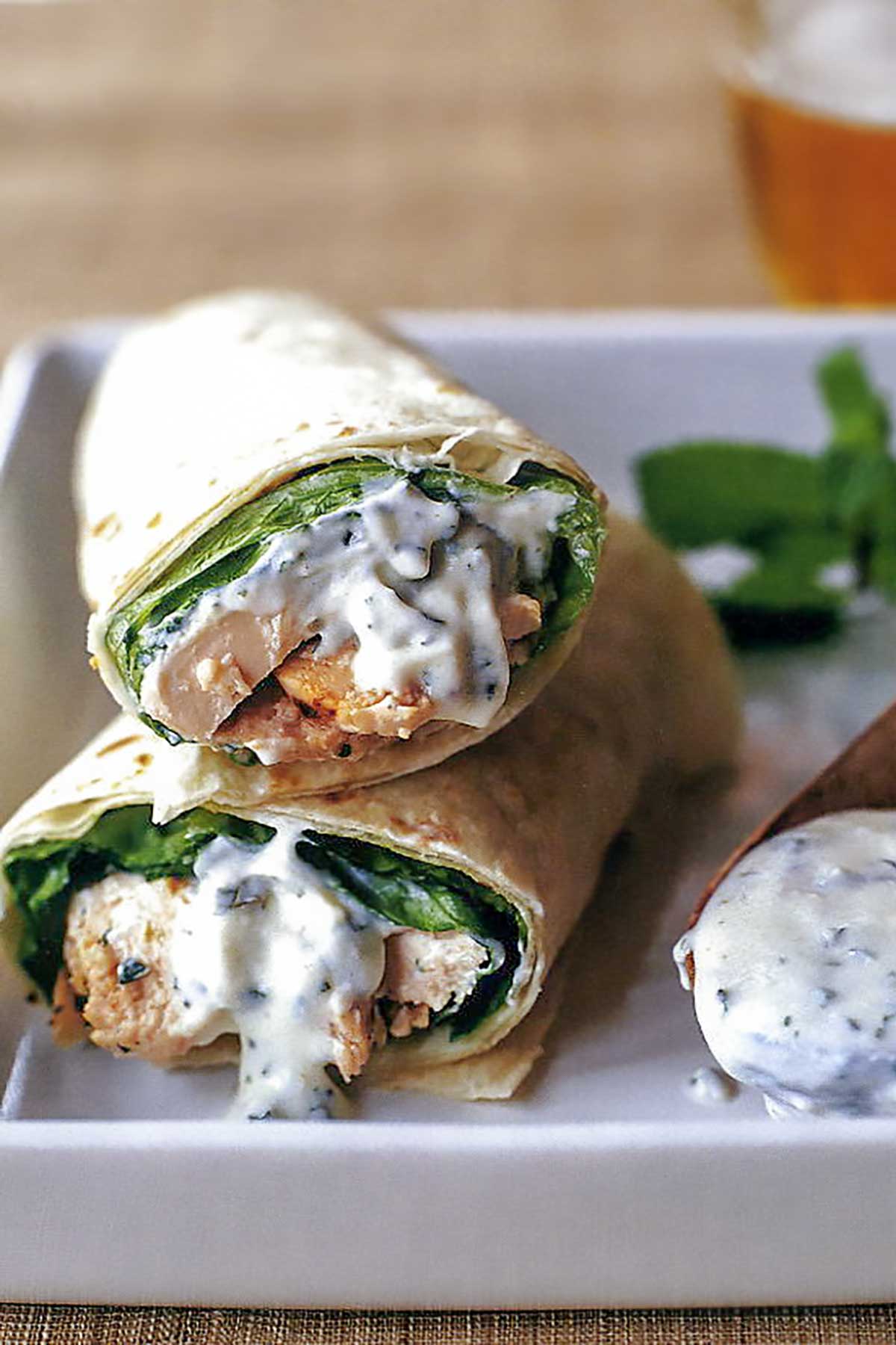 A tandoori chicken wrap sliced in half with yogurt sauce dripping out and a wooden spoon with yogurt sauce on the side.