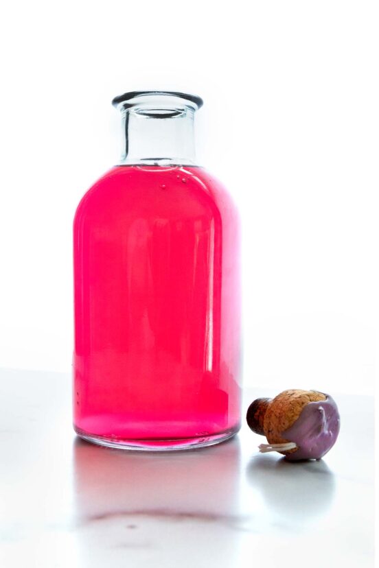 A clear glass bottle filled with vibrant, blush-pink homemade chive blossom vinegar set against a bright, minimalist background.