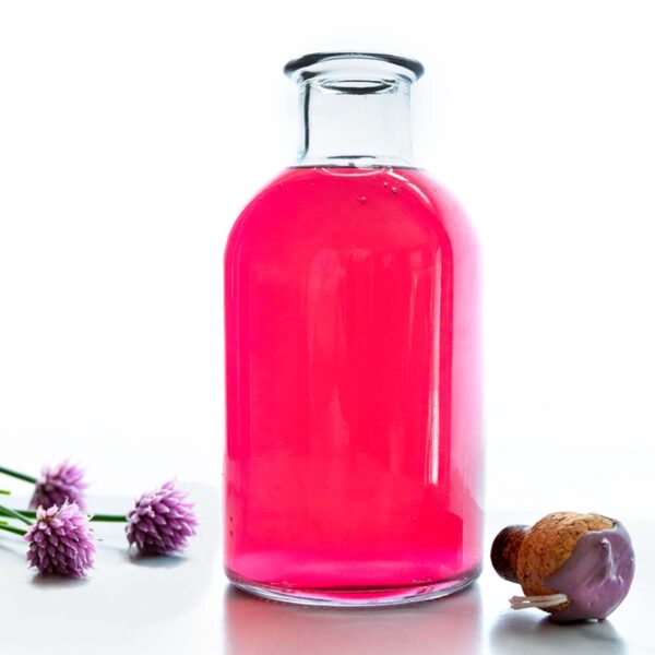 An apothecary jar of magenta-pink chive blossom vinegar; a cork and chive blossoms rest nearby.