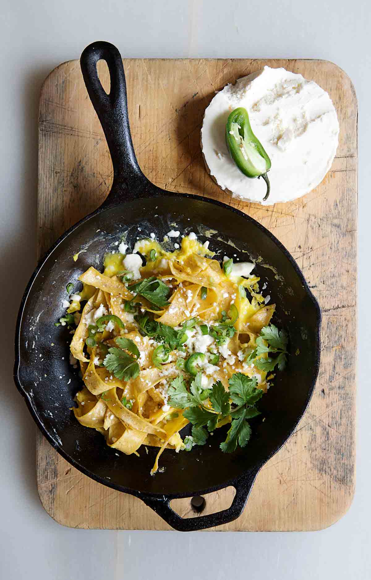 A skillet of chilaquiles on a wooden board with half a jalapeno and block of cotija cheese on the side.