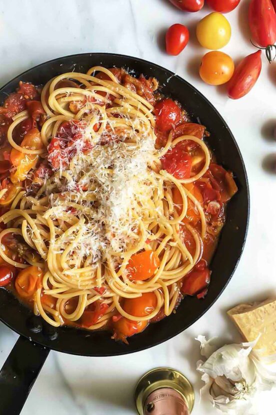 A skillet filled with spaghetti and cherry tomatoes, with Parmesan, garlic, and tomatoes scattered around it.