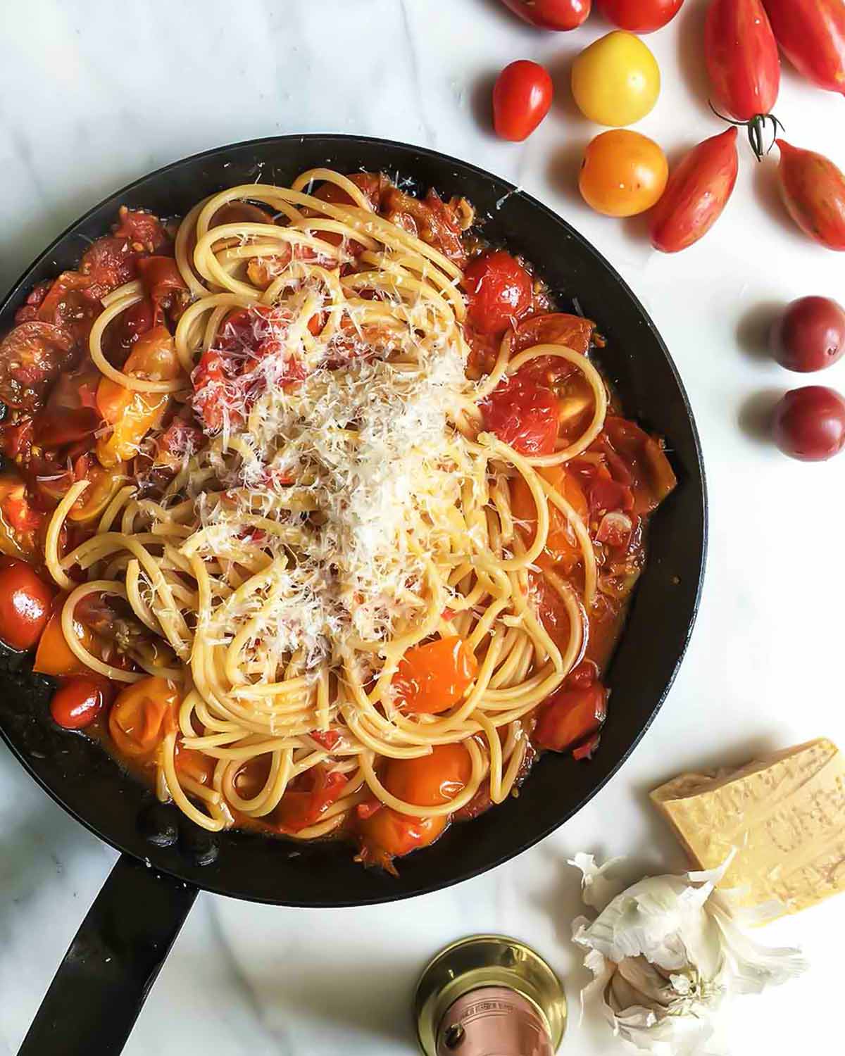 A skillet filled with spaghetti and cherry tomatoes, with Parmesan, garlic, and tomatoes scattered around it.