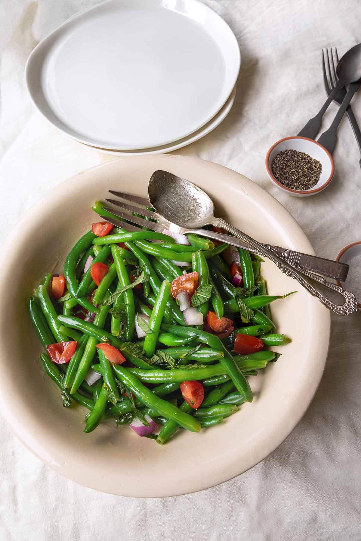 A bowl of green bean salad with mint and tomatoes with two plates, utensils, and a dish of pepper in the background.
