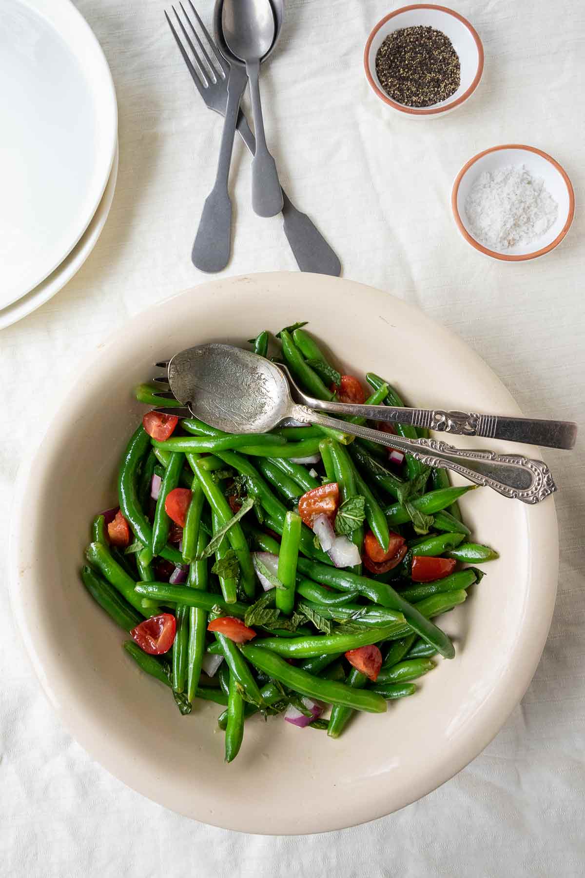 A bowl of green bean salad with mint and tomatoes with two plates, utensils, and a dish of pepper in the background.