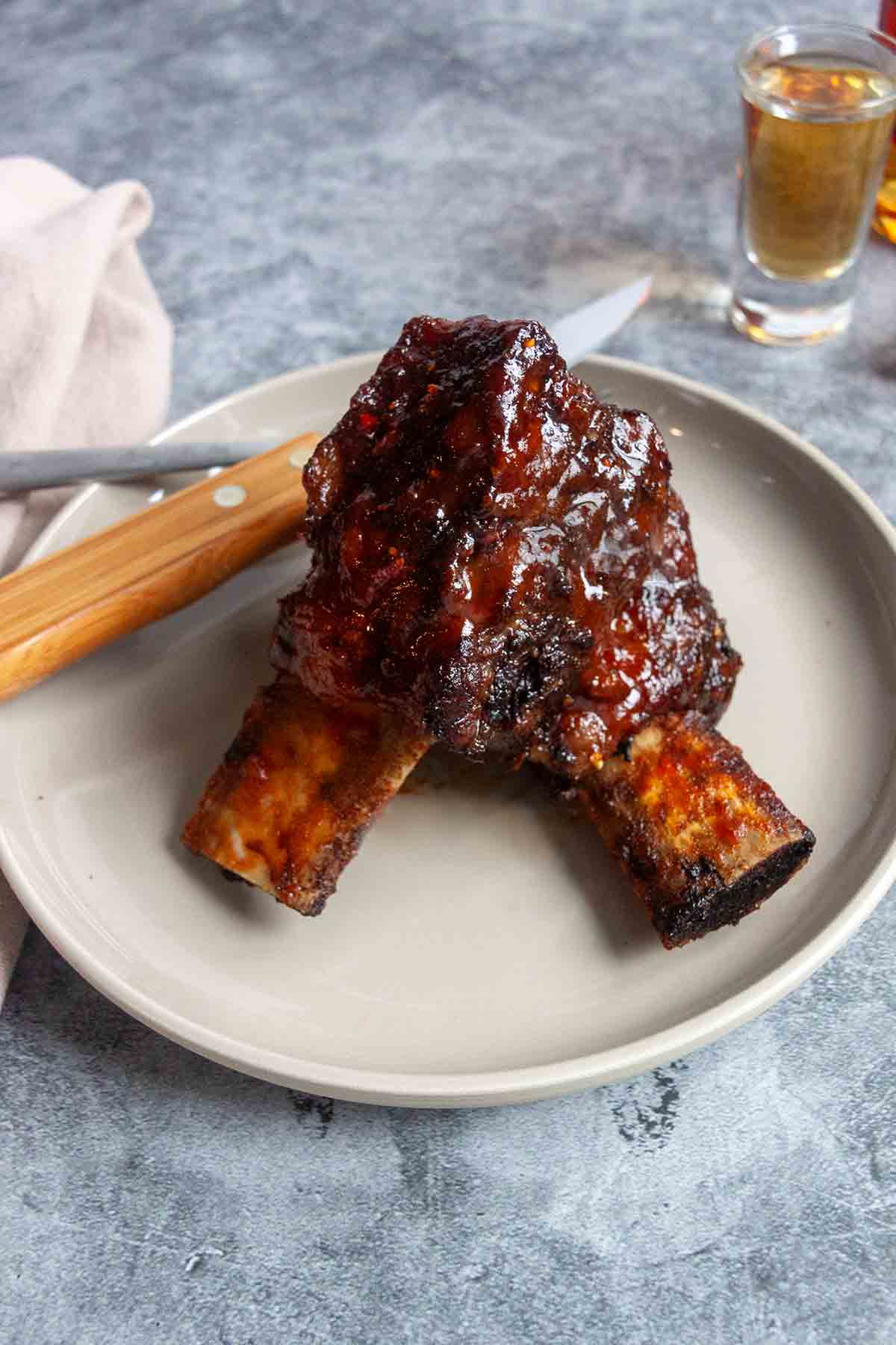 Two cooked short ribs on a plate with a shot of bourbon in the background and a fork and knife on the side.