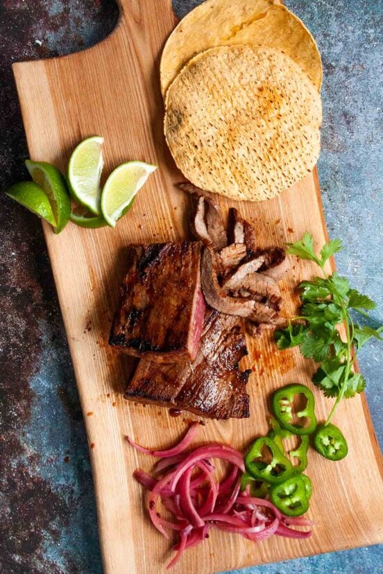 Sliced skirt steak on a wooden board with tortillas, lime wedges, cilantro, jalapeno, and pickled red onion.