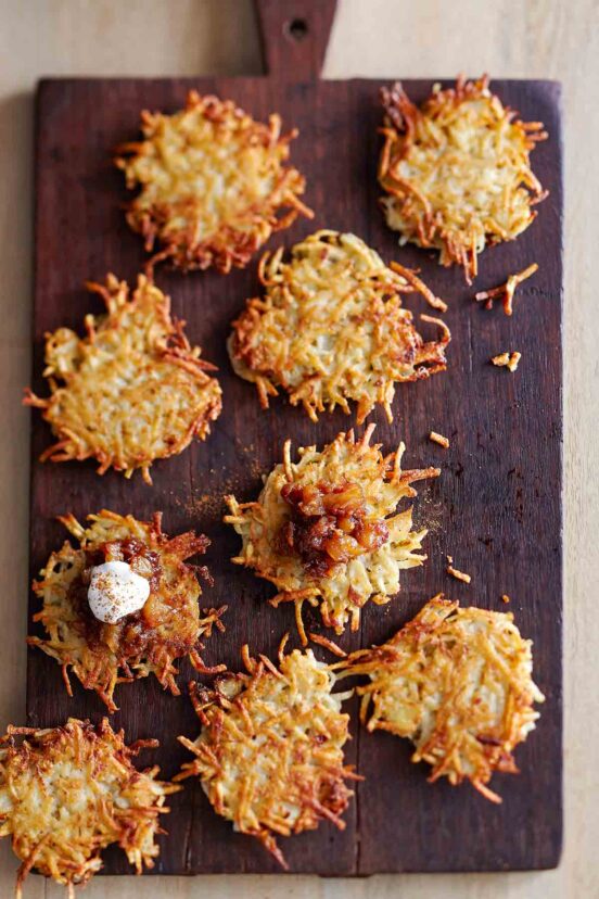 Nine potato latkes on a wooden board, with a dollop of chutney on two of them and sour cream on one of them.