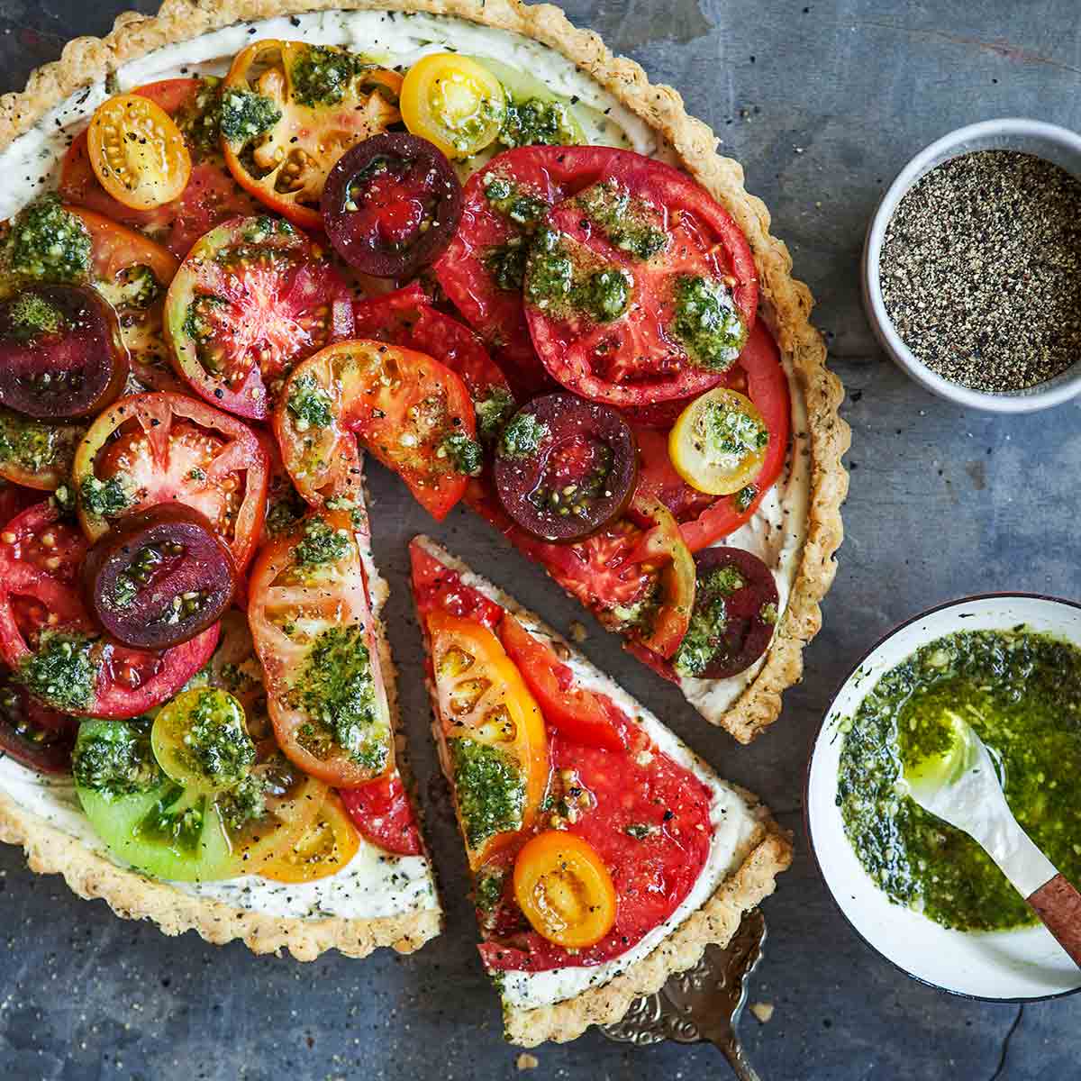 A fresh tomato tart with one slice being lifted from it and a bowl of pesto and dish of pepper on the side.