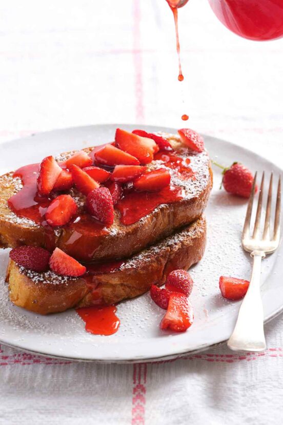 Strawberry syrup being poured onto two slices of French toast topped with sliced strawberries.