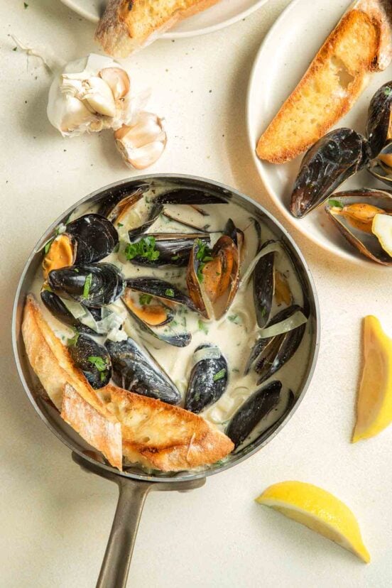 A copper saucepan filled with mussels in a creamy white wine garlic sauce, slices of toasted bread. Nearby, a head of garlic and lemon slices.