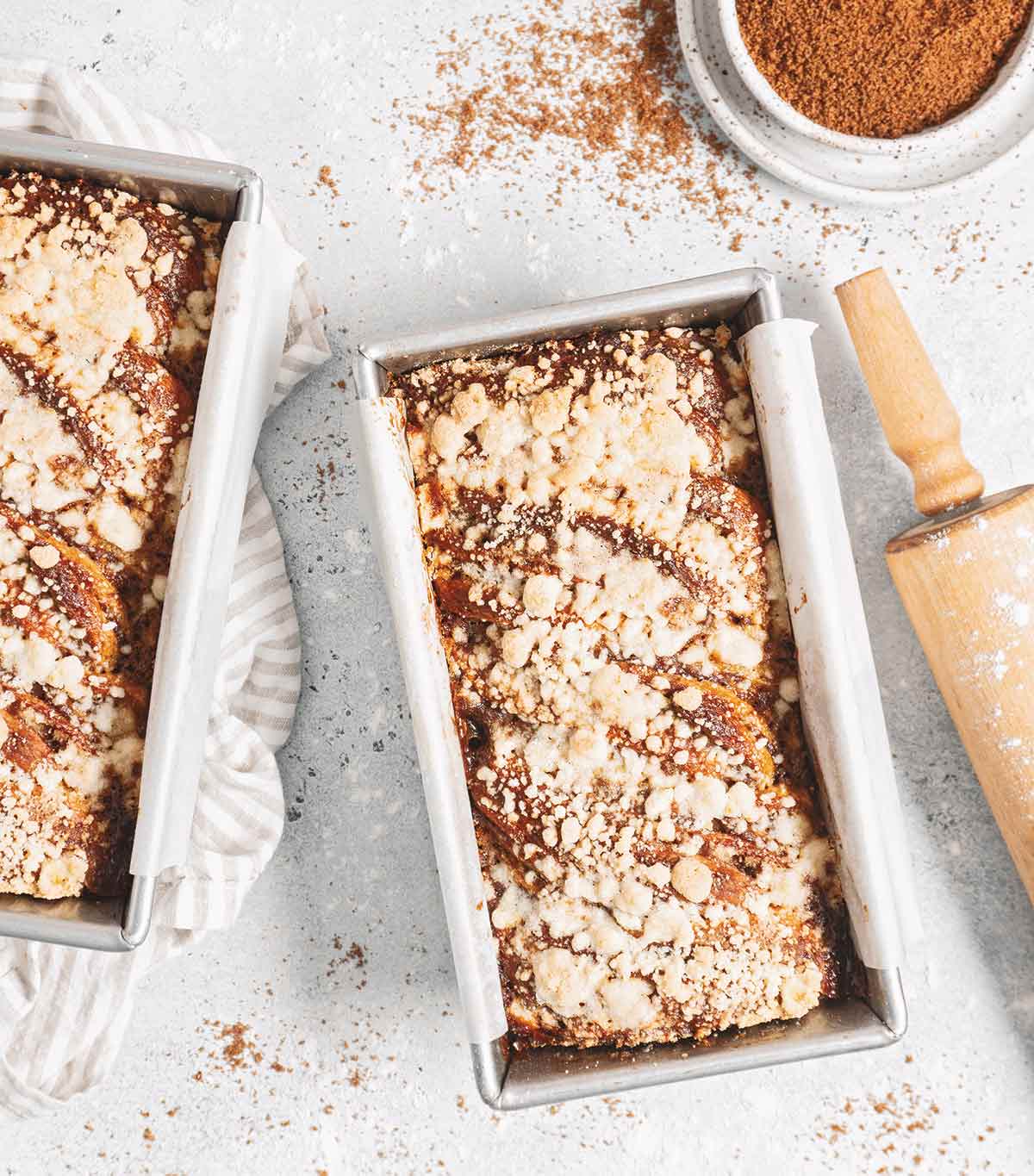 Two loaves of pumpkin pie babka in loaf tins with a rolling pin and a dish of brown sugar on the side.