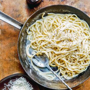 A skillet filled with cacio e pepe, with a bowl of shredded Parmesan cheese on the side.