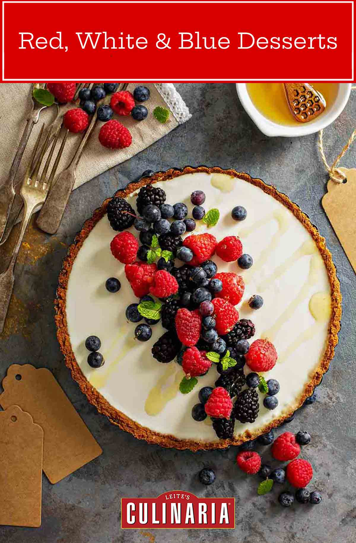 A cheesecake tart with a graham cracker crust and assorted fresh berries on top, a dish of honey, and some berries and forks around it.