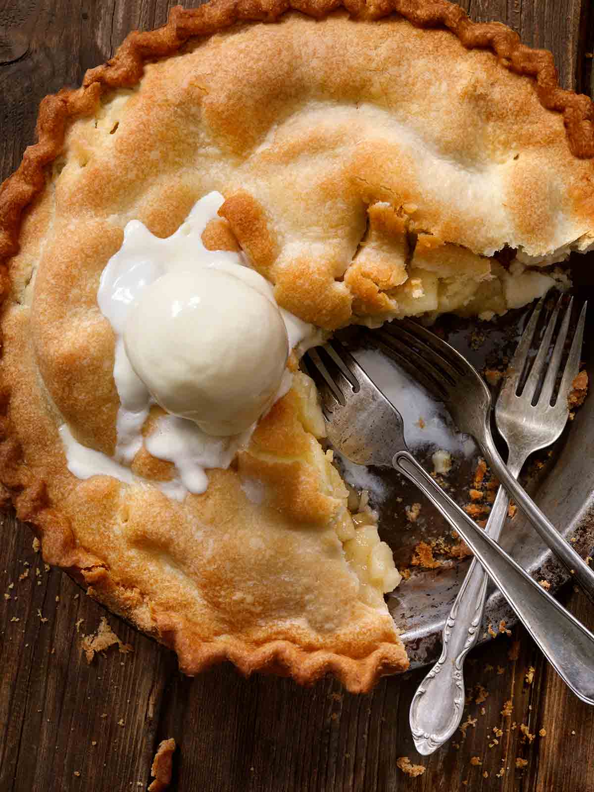 A classic apple pie with a scoop of melting ice cream on top, one slice missing, and three forks resting in the pie dish.
