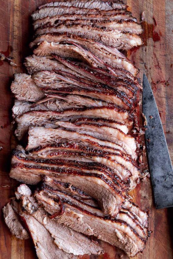 A sliced brisket on a wooden board with a carving knife resting beside it.