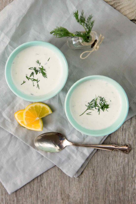 Two bowls of cold cucumber soup with a spoon, orange slices, and dill sprigs on the side.
