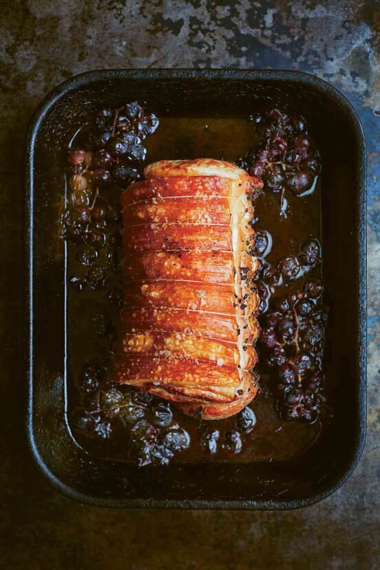 A roast pork loin in a black roasting pan surrounded by a crushed grape sauce.