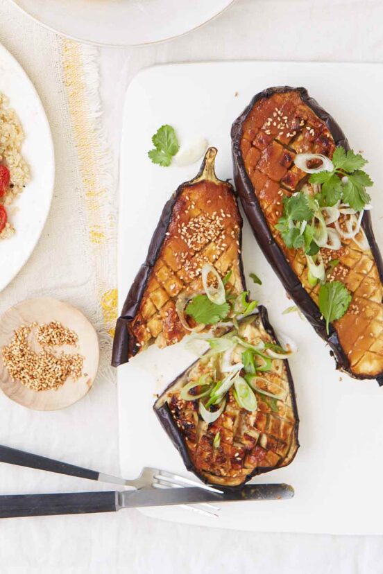 A miso-glazed eggplant on a white platter, topped with scallions and cilantro, with a dish of sesame seeds on the side.