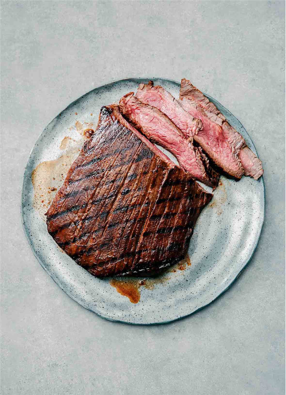 A piece of grilled flank steak on a plate with a few slices cut from it.
