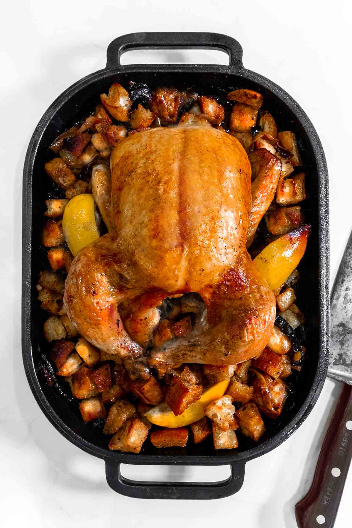 A roast lemon chicken on a bed of croutons in a metal roaster with a knife on the side.