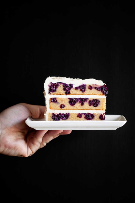A person holding a slice of frosted three layer blueberry lemon cake on a white plate.