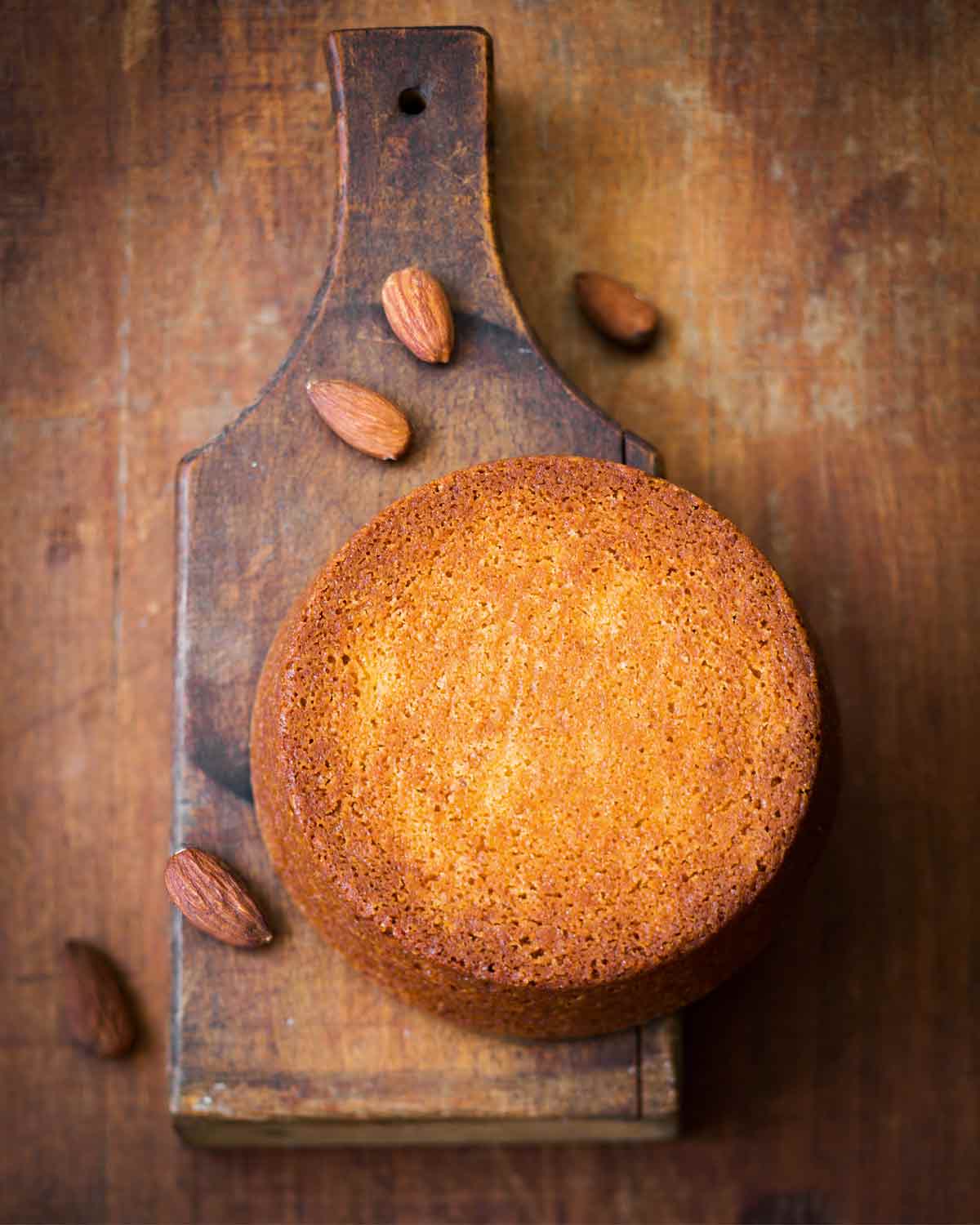 A flourless almond cake on a wooden cutting board with some almonds scattered around it.