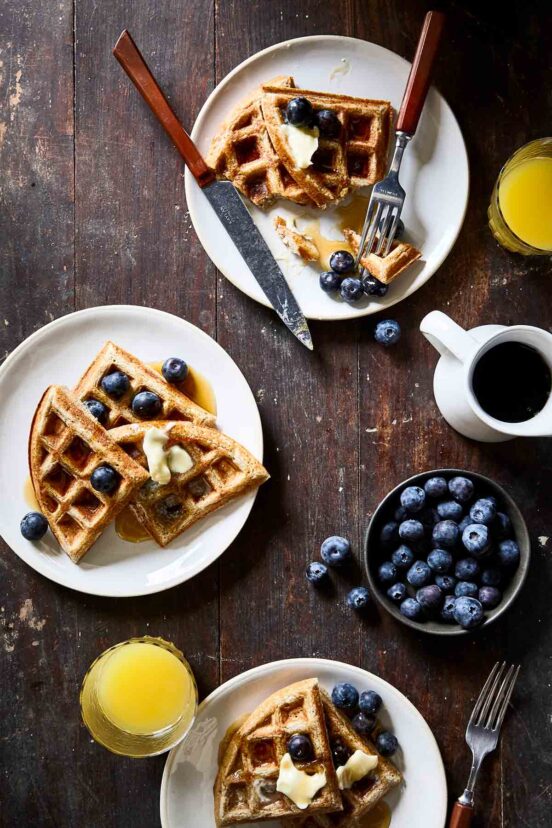 Three plates topped with waffles, butter, and blueberries, with a dish of blueberries, a pitchers of syrup and a couple glasses of orange juice on the side.