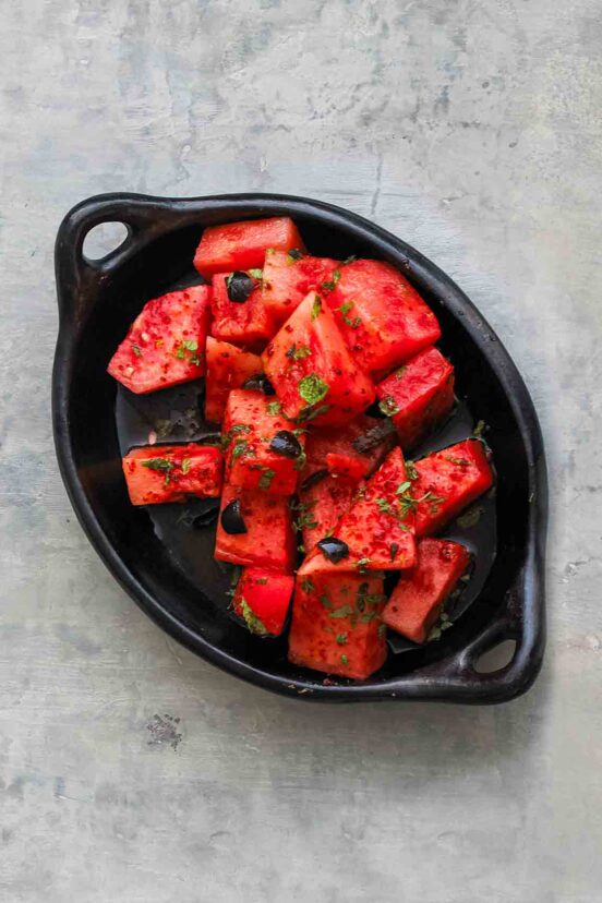 A black dish filled with cubes of watermelon that is topped with olives, mint, and aleppo pepper.