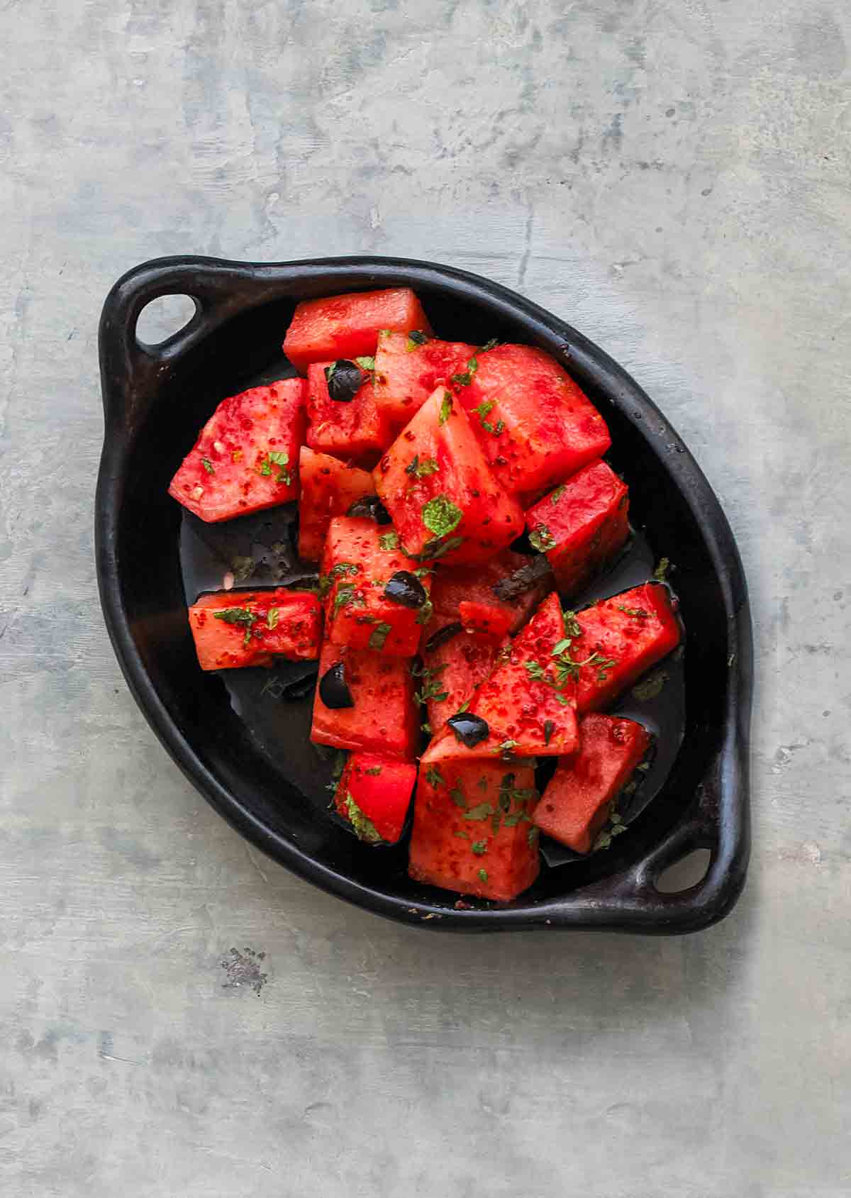 A black dish filled with cubes of watermelon that is topped with olives, mint, and aleppo pepper.