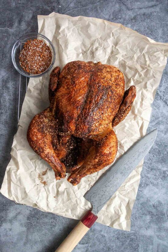 A smoked chicken on a piece of crumpled parchment paper with a knife and a bowl of spice rub on the side.