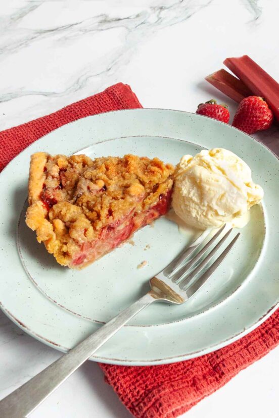 A slice of strawberry rhubarb pie on a green plate with a scoop of ice cream and a fork on the side.