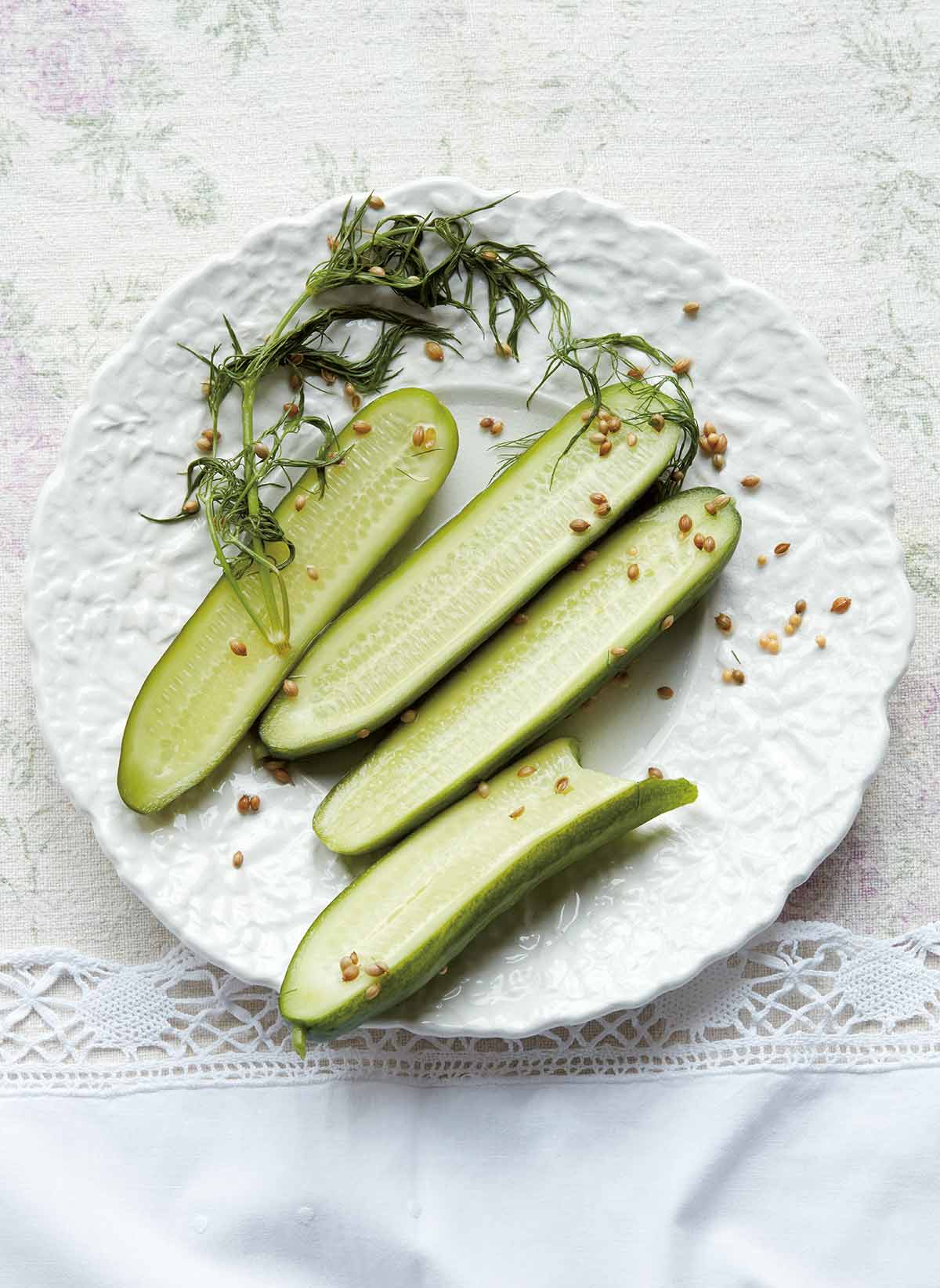 Four halved quick dill pickles with a sprig of dill and some seeds on a white plate.