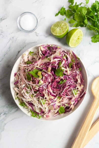 A bowl of apple coleslaw with wooden serving spoons, salt, two lime halves, and some cilantro on the side.