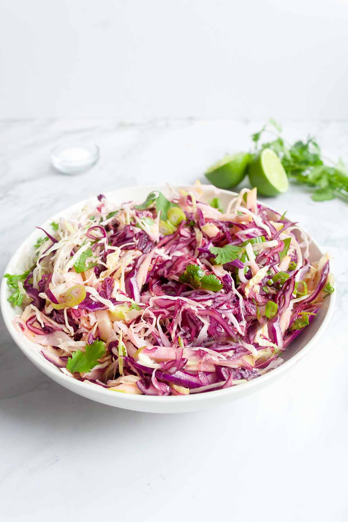 A bowl of apple coleslaw with wooden serving spoons, salt, two lime halves, and some cilantro on the side.