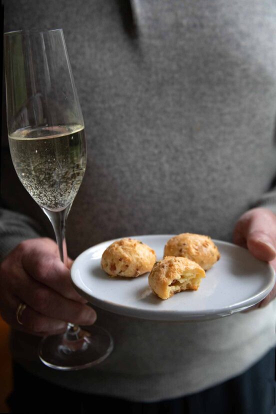 A person holding a glass of Champagne and a plate with three prosciutto cheese gougeres.