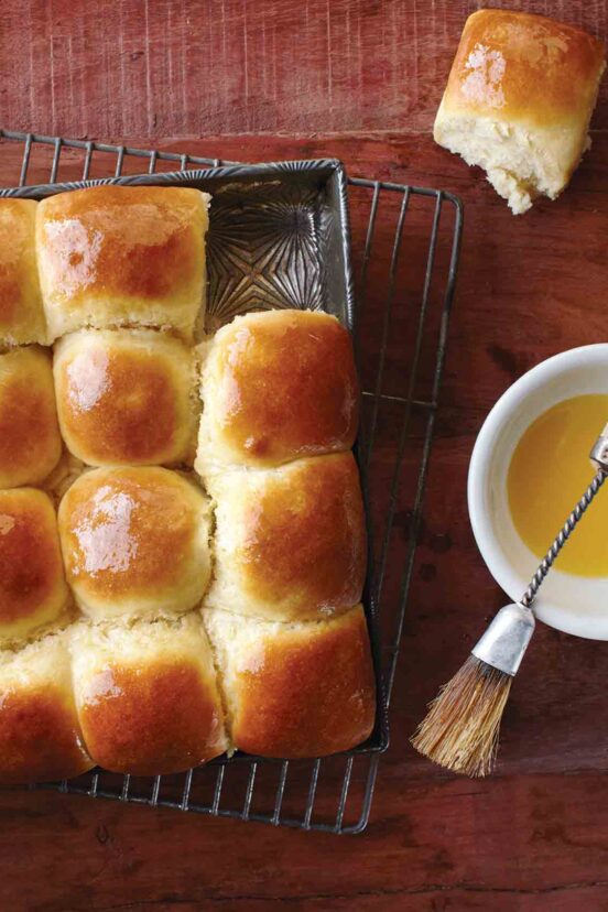A tray of dinner rolls with one removed and a dish of butter and brush on the side.