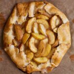 A white peach crostata--slices of white peaches in a circle of dough folded over on the edges sitting a sheet of parchment.