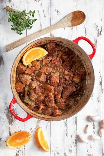A red Dutch oven filled with braised pork cubes with an orange wedge on top and a few orange wedges, garlic cloves, oregano, and spoon on the side.