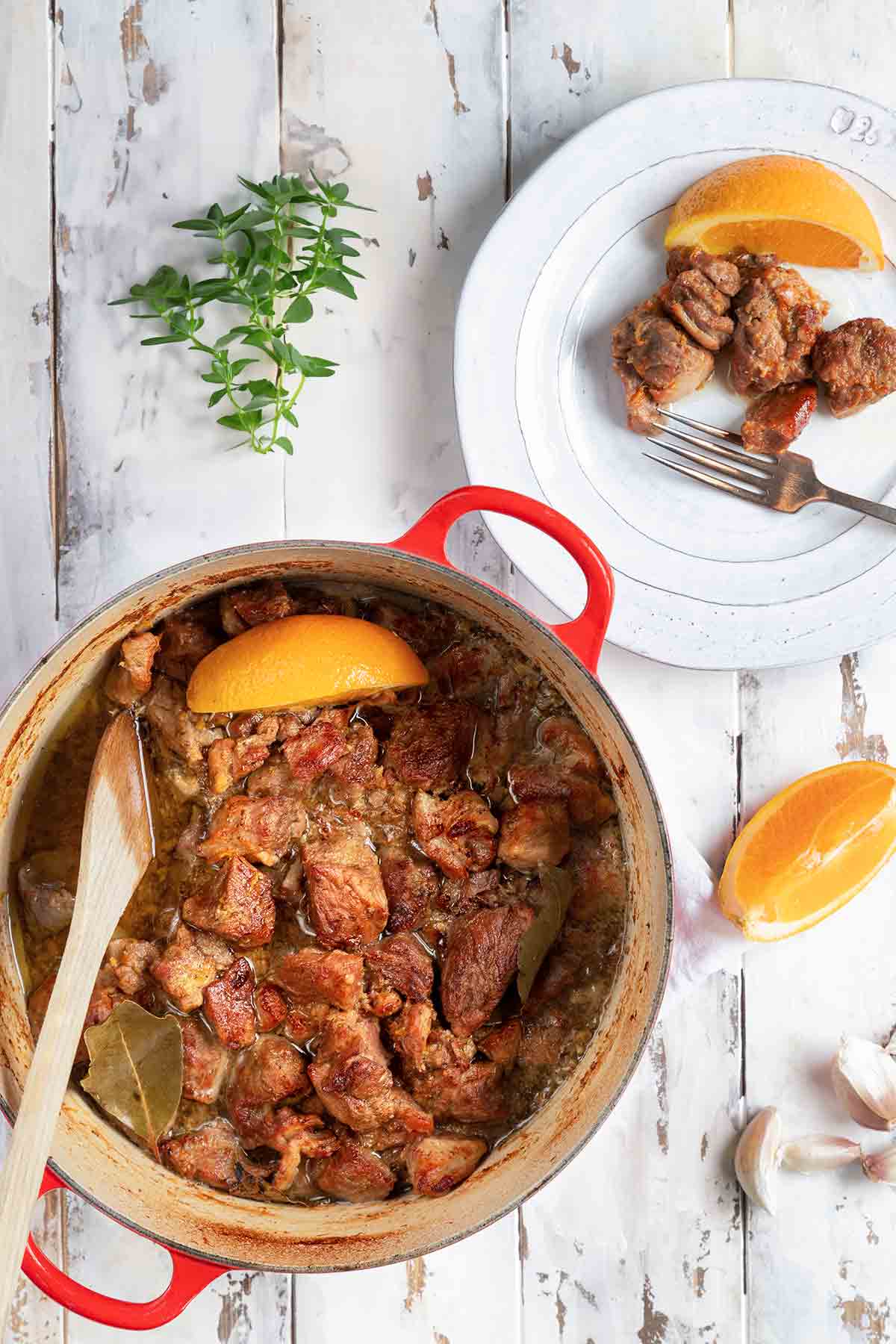 A red Dutch oven filled with braised pork cubes with an orange wedge on top and a few orange wedges, garlic cloves, oregano, and spoon on the side.