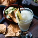 A tall glass of frozen margarita with a salt rim and lime wheel beside a bowl of chips and guacamole.
