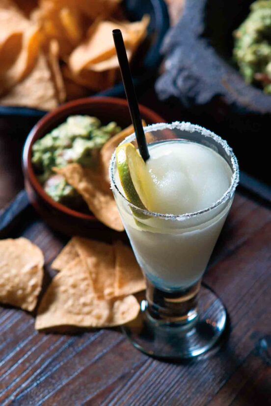 A tall glass of frozen margarita with a salt rim and lime wheel beside a bowl of chips and guacamole.