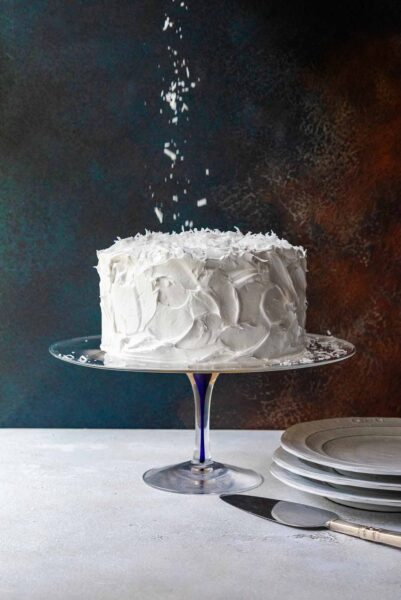 A white old-fashioned coconut cake with 7-minute frosting topped sprinkled with shredded coconut on a glass cake stand.