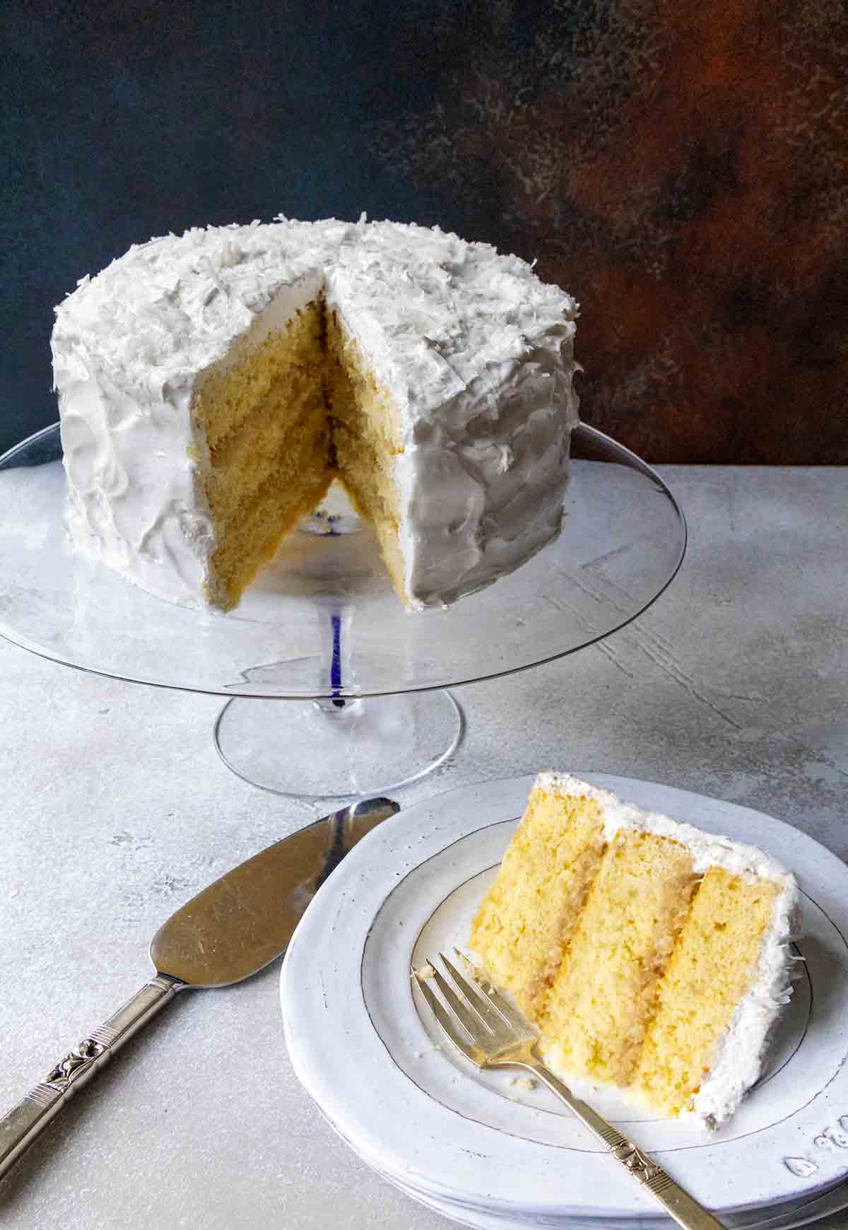 A white old-fashioned coconut cake with 7-minute frosting topped with shredded coconut on a glass cake stand. A slice in front.