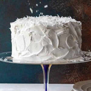 A white old-fashioned coconut cake with 7-minute frosting topped sprinkled with shredded coconut on a glass cake stand.